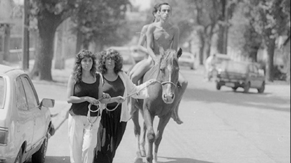 Dos hombres desnudos sobre un cabello llevado por una cuerda por dos mujeres en una calle.