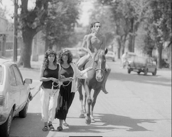 Dos hombres desnudos sobre un cabello llevado por una cuerda por dos mujeres en una calle.